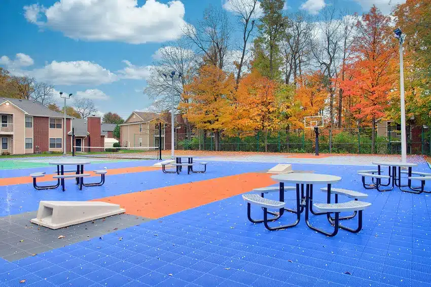 Outdoor sports court with blue and orange flooring, picnic tables, and a basketball hoop, surrounded by trees with autumn foliage and residential buildings in the background.