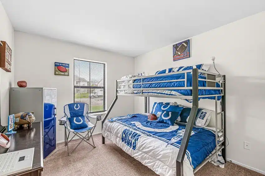 A bedroom with a bunk bed, blue football-themed bedding, folding chair, small desk, and window letting in natural light. Sports decor and memorabilia are displayed on the walls.