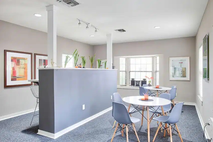 Modern room with blue chairs around a round table, a half-wall divider with bar stools, framed artwork on walls, and several potted plants on the divider.