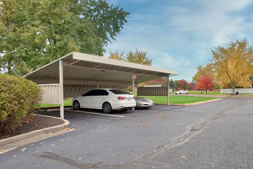 Two cars are parked under a metal carport in a paved parking lot surrounded by trees and shrubs on a partly cloudy day.