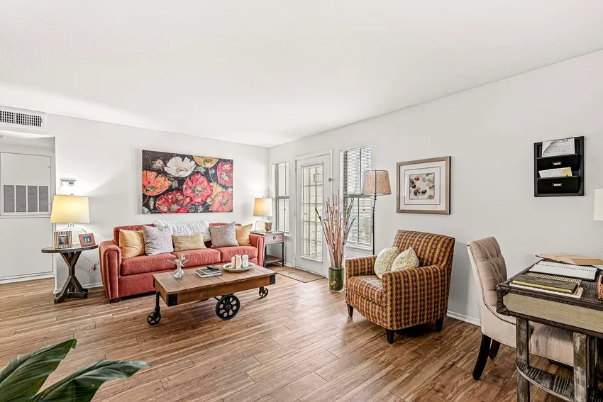 A bright living room with wood flooring, a red sofa, patterned armchair, coffee table, desk, and floral artwork on the wall.