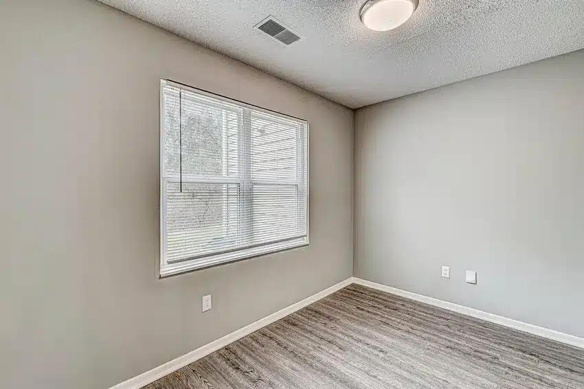 Empty room with light gray walls, a large window with blinds, wood-style flooring, and a ceiling light fixture.