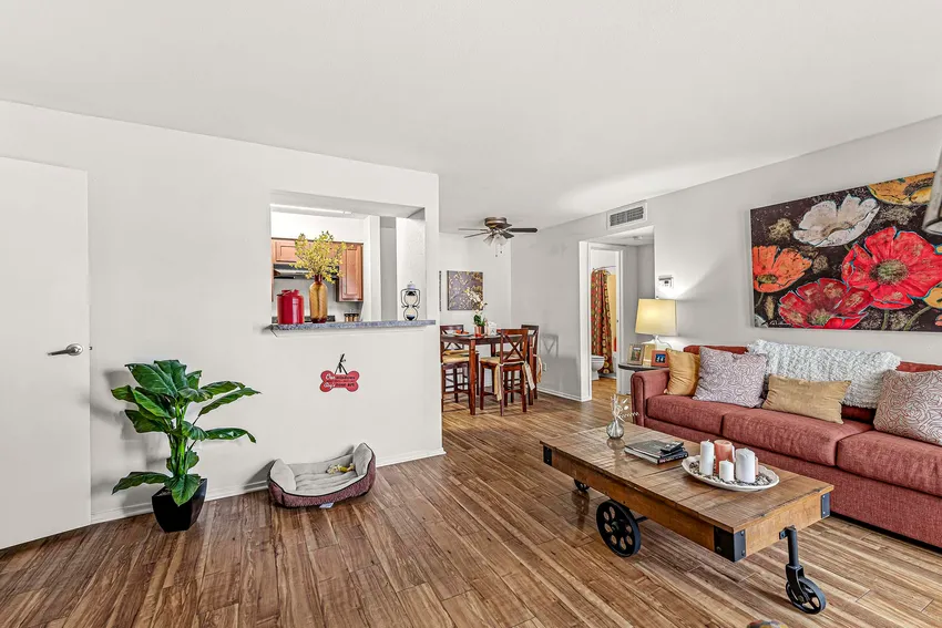 A living room with wood flooring, a red sofa, a coffee table on wheels, a potted plant, a pet bed, and wall art; a dining area and kitchen are visible in the background.