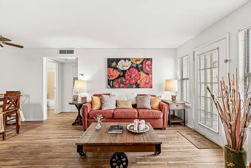 A living room with a red sofa, floral wall art, wooden coffee table, two table lamps, and large windows letting in natural light. Dining area visible in the background.