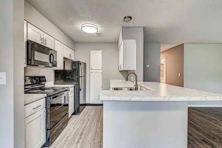 Modern kitchen with white cabinets, black appliances, marble-patterned countertops, and wood-style flooring, adjacent to a partially visible open living area.