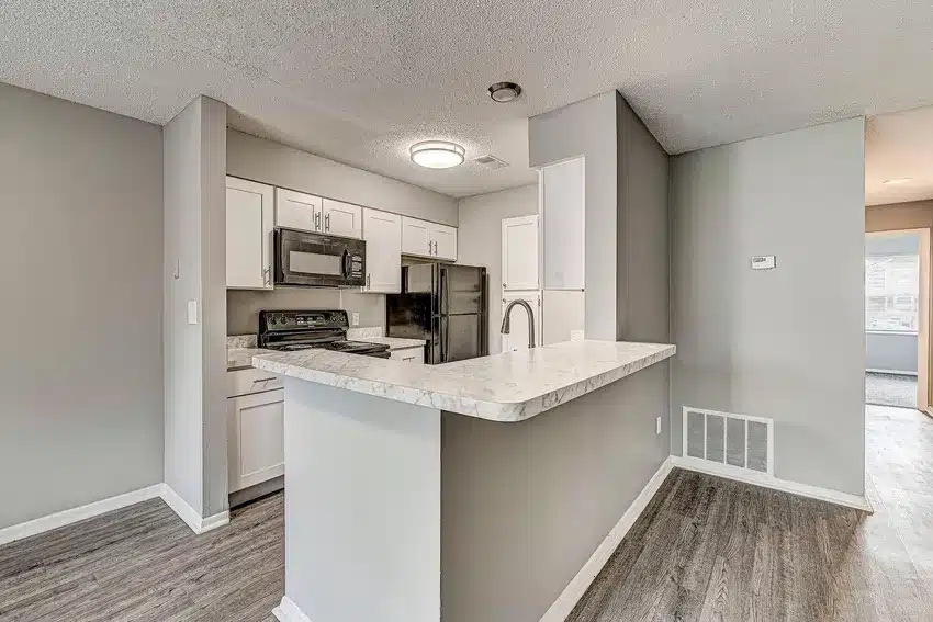 Modern kitchen with white cabinets, stainless steel appliances, marble countertop, and grey walls, adjacent to an open living area with wood-style flooring.