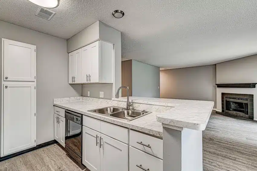 Modern kitchen with white cabinets, marble countertops, double sink, dishwasher, and an open view into a living area with a fireplace.