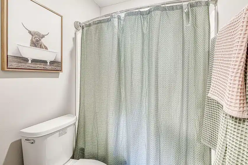 A bathroom with a toilet, a bathtub behind a patterned shower curtain, towels hanging on the wall, and framed artwork of a cow in a bathtub.