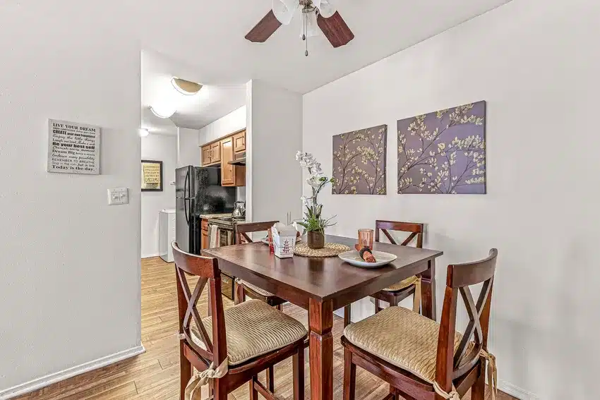 A small dining area with a square wooden table and four chairs, set for two, adjacent to a kitchen with wood cabinets and black appliances. Two floral paintings hang on the wall.