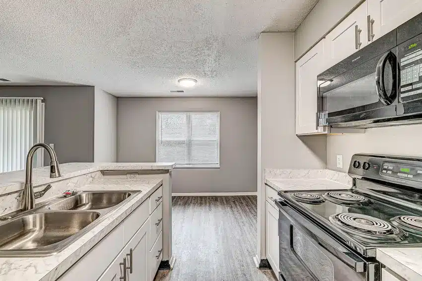 Modern kitchen with white cabinets, stainless steel double sink, electric stove, black microwave, and a view into a dining area with a window and wood-style flooring.