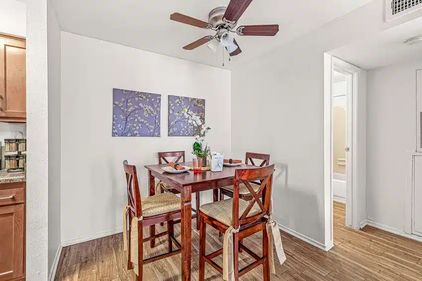 A small dining area with a wooden table and four chairs, a ceiling fan above, two floral paintings on the wall, and a view into a bathroom and kitchen.