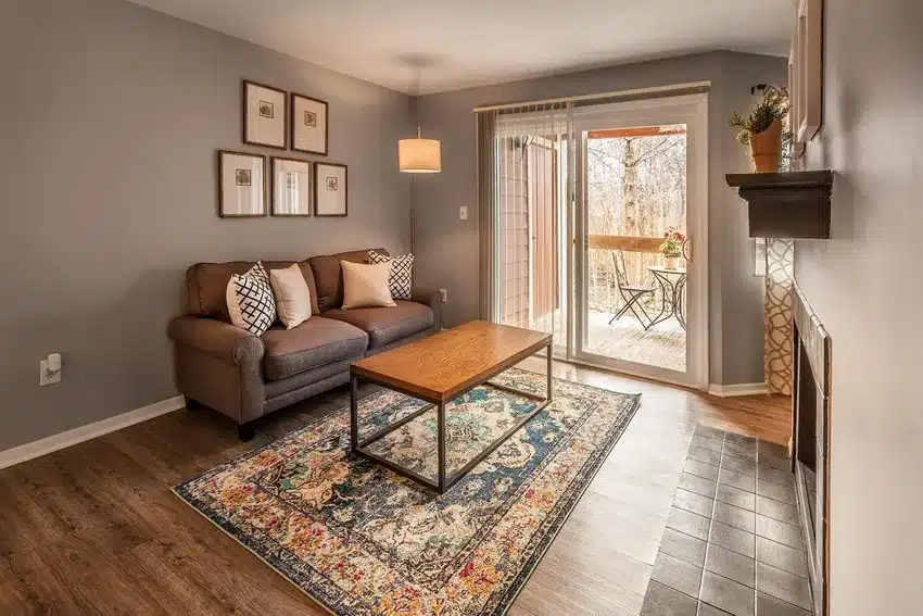 A living room with a gray sofa, wooden coffee table, patterned rug, wall art, and sliding glass doors leading to a balcony with outdoor seating.