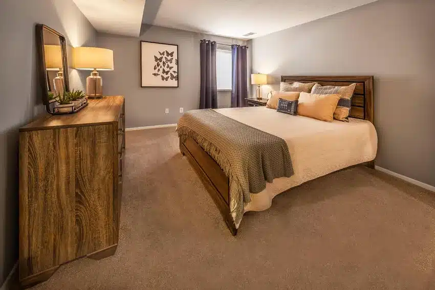 A bedroom with a wooden bed, beige bedding, matching dresser with lamps, carpeted floor, framed artwork on the wall, and a window with dark curtains.