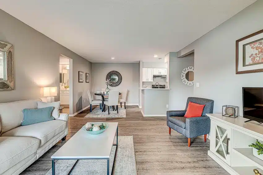 Modern living room with a beige sofa, coffee table, armchair, TV stand, and dining area; neutral colors and wood flooring throughout the open space.