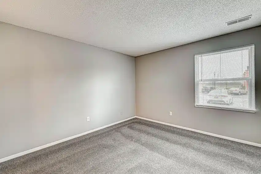 Empty room with light gray carpet, beige walls, and a window with blinds letting in daylight; an electrical outlet and vent are visible.