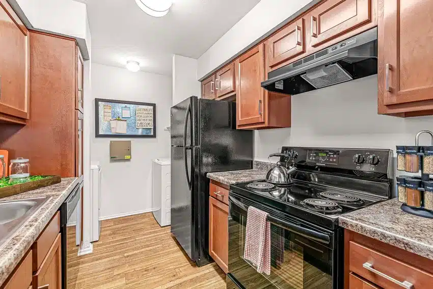 A modern galley kitchen features black appliances, brown cabinets, marble-look countertops, and hardwood floors, with a washer and dryer visible in an adjacent laundry area.