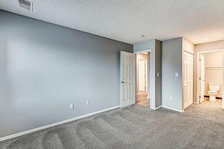 Empty bedroom with gray carpet, light gray walls, an open door leading to a hallway, and a closet with double doors. A bathroom with visible toilet is in the background.