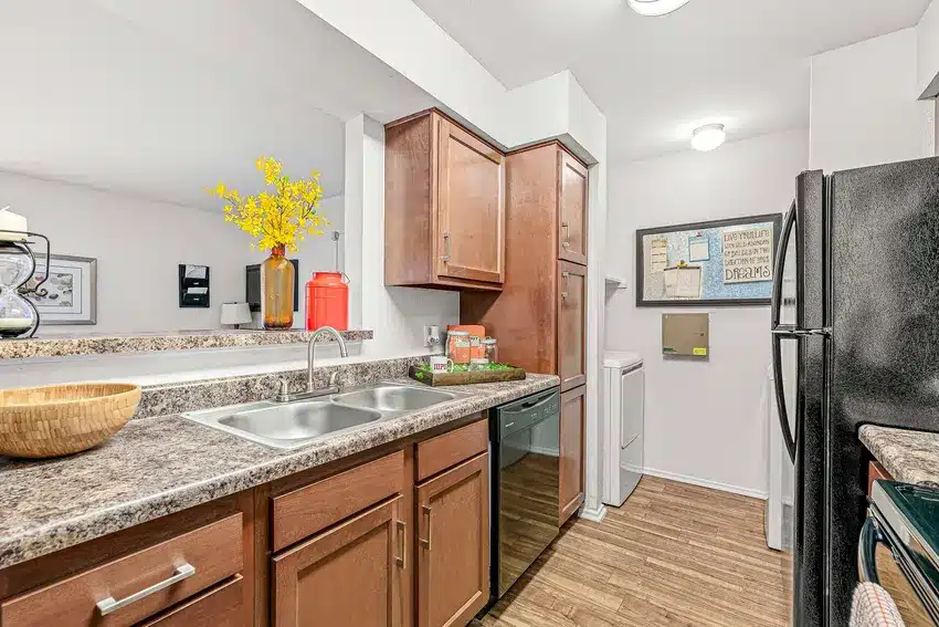 Modern kitchen with brown cabinets, marble countertops, double sink, and black appliances; laundry area with washer and dryer visible in the background.