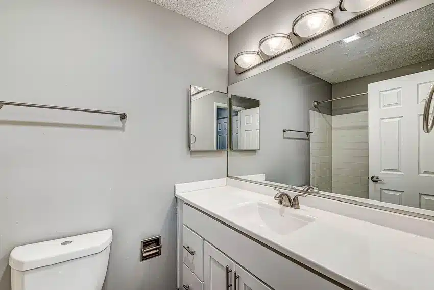 Modern bathroom with a white vanity, single sink, wall-mounted mirror, overhead lights, towel rack, and toilet against light gray walls.