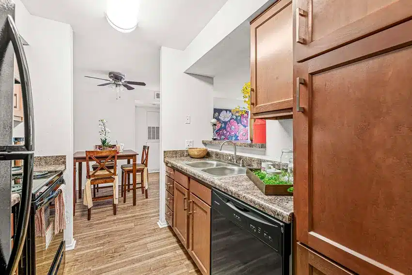 A narrow kitchen with brown cabinets, granite countertops, a double sink, and a dishwasher opens to a dining area with a wooden table and chairs.