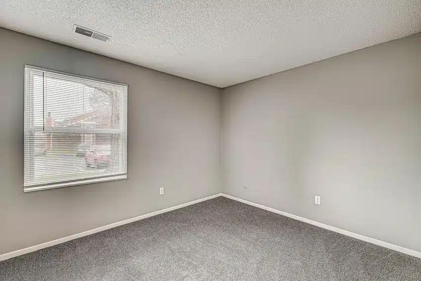 Empty room with gray carpet, light gray walls, a window with blinds, and a ceiling vent. Natural light enters through the window.
