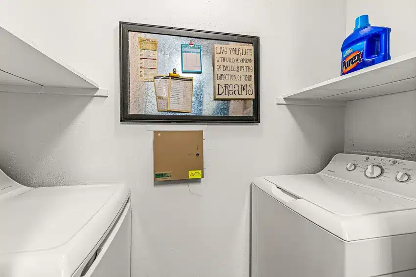 A laundry room with a washing machine, a dryer, shelves, a detergent bottle, and a framed wall organizer with notes and quotes.