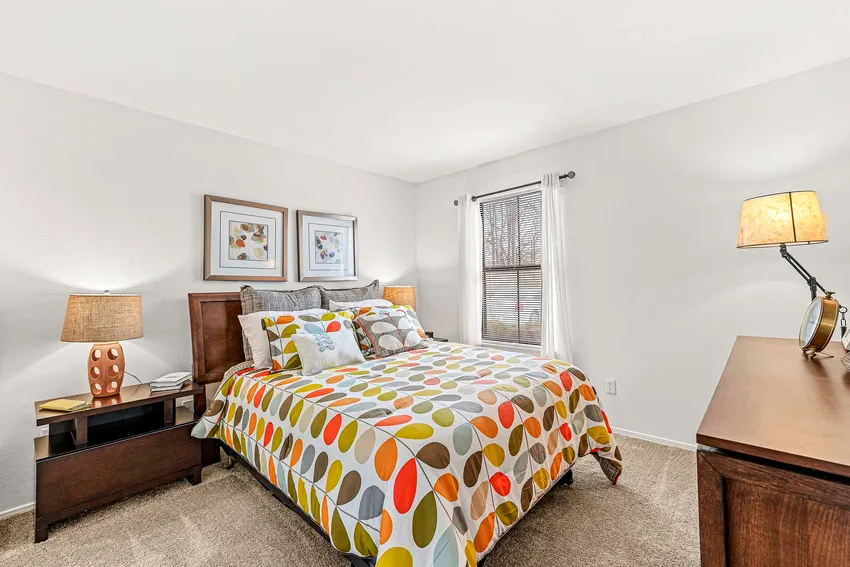 A neatly made bed with a colorful patterned comforter sits in a bright bedroom with a nightstand, lamps, dresser, wall art, and a window with white curtains.