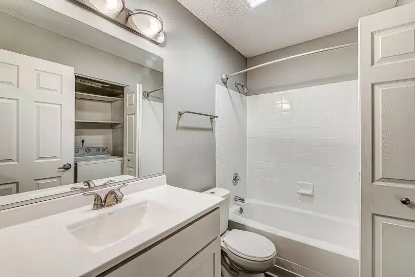 A bathroom with a white vanity, sink, and toilet, a bathtub with a shower curtain rod, and an open door showing a washer and dryer in an adjacent room.