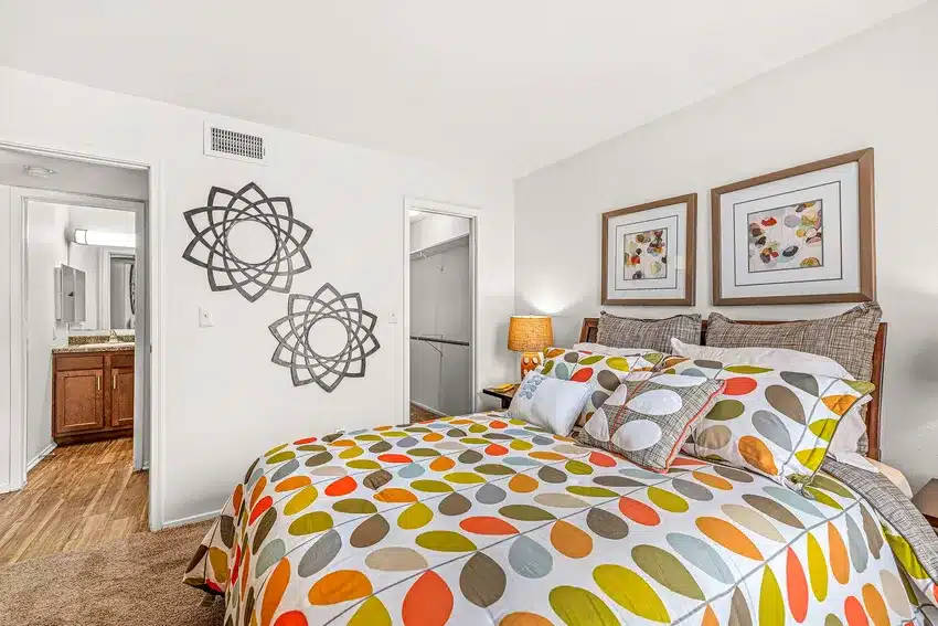 A bedroom with a patterned bedspread, decorative pillows, two framed pictures above the bed, and modern wall art; open doors reveal a closet and bathroom.