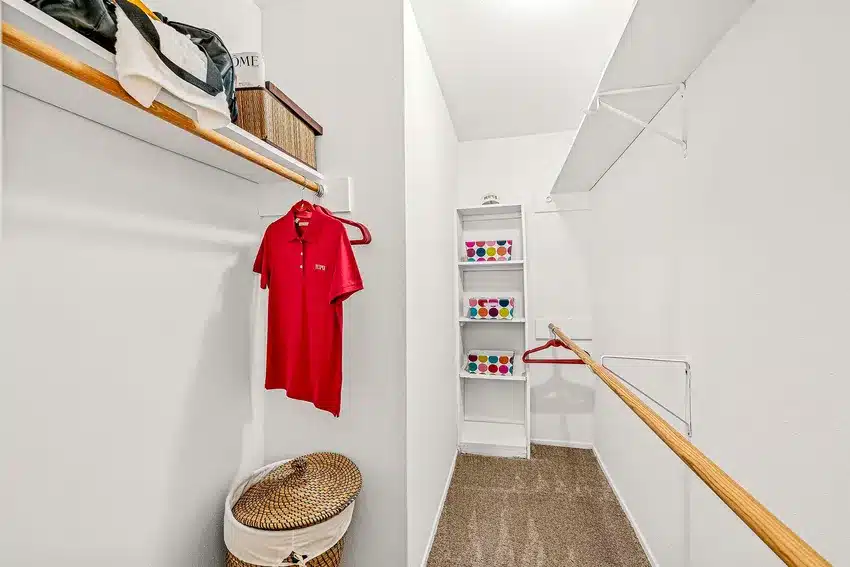 A walk-in closet with hanging rods, a single red shirt on a hanger, shelves with colorful boxes, a wicker hamper, and carpeted flooring.