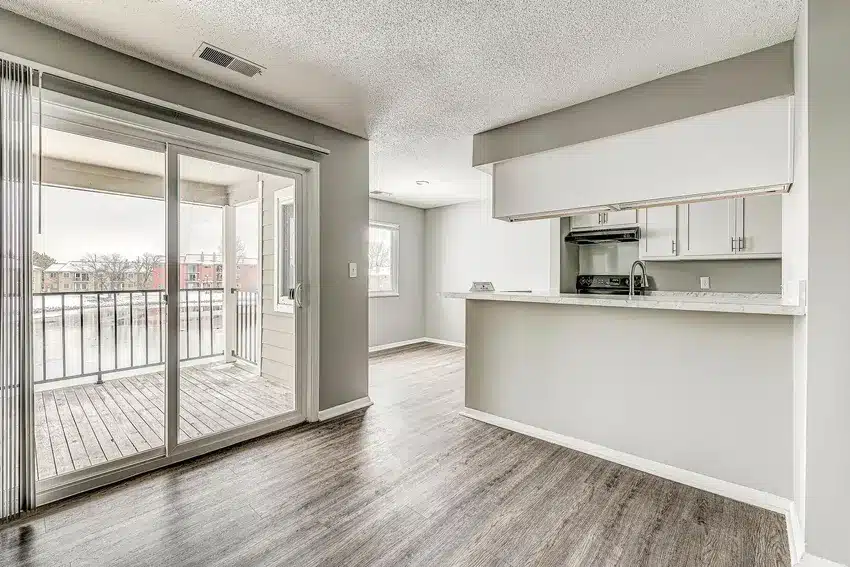 A modern apartment interior with wood flooring, an open kitchen, and sliding glass doors leading to a covered balcony.