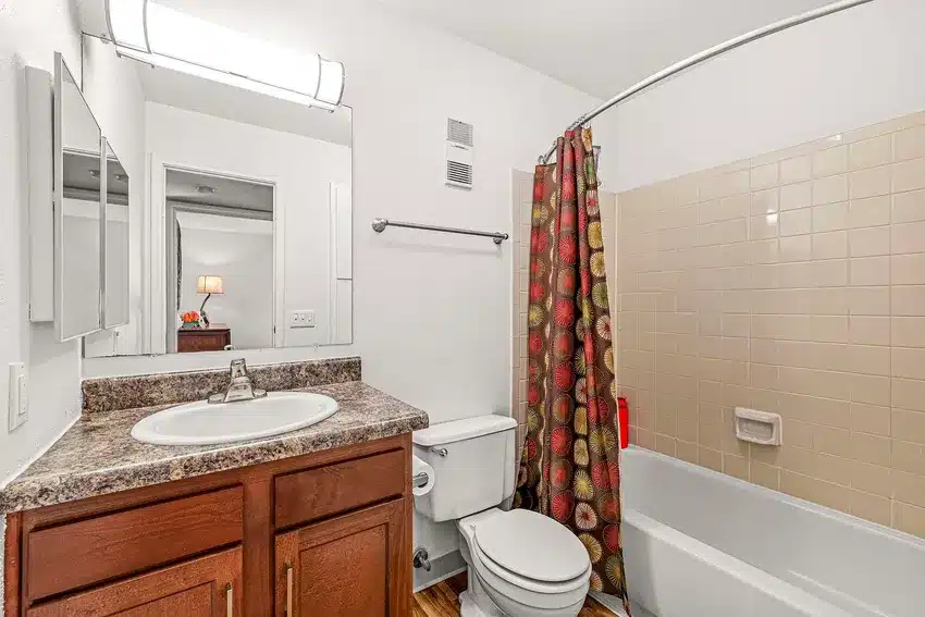 A bathroom with a sink, mirror, toilet, and bathtub with beige tiles and a patterned shower curtain; a bedroom is visible through the open door.