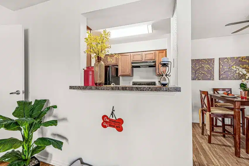 Open kitchen view with brown cabinets, countertop decor including vases and a lantern, a dining area with wooden chairs, and a plant in the foreground.
