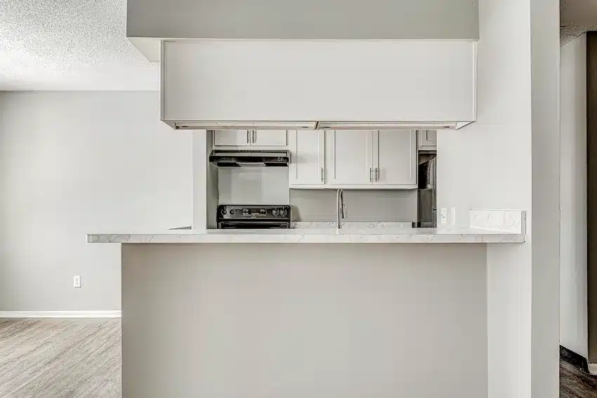 A modern kitchen with white cabinets, a marble countertop, and stainless steel appliances, viewed from across a breakfast bar.