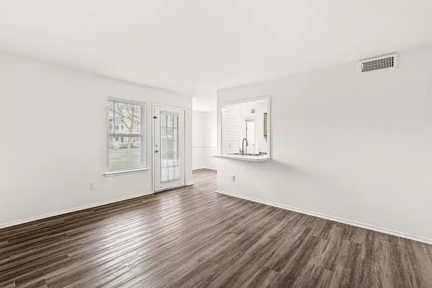 Bright, empty living room with wood flooring, white walls, a window and glass door on the left, and an open kitchen pass-through on the right.