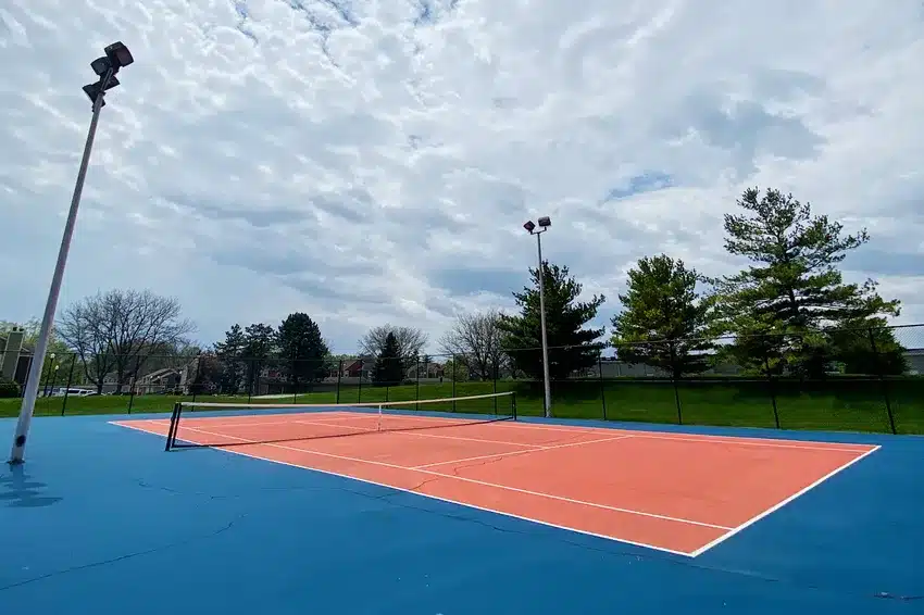 An empty outdoor tennis court with a blue surface and orange playing area, surrounded by a fence and trees under a partly cloudy sky.