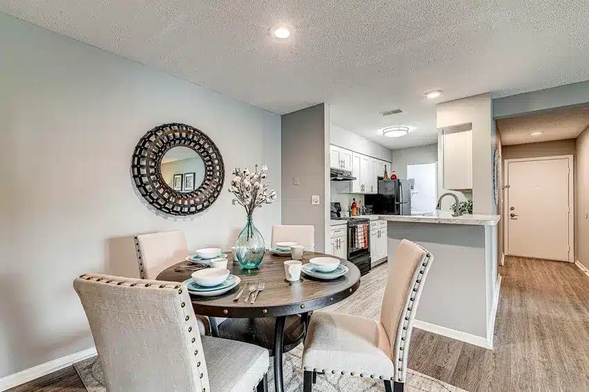 Modern dining area with round table set for four, upholstered chairs, decorative mirror on wall, and view into a white kitchen with stainless steel appliances.