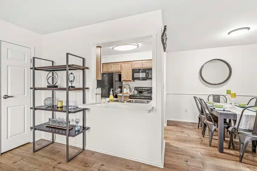 Open kitchen with wood cabinets and black appliances, adjacent to dining area with metal chairs and a round mirror, and a decorative shelving unit in the foreground.