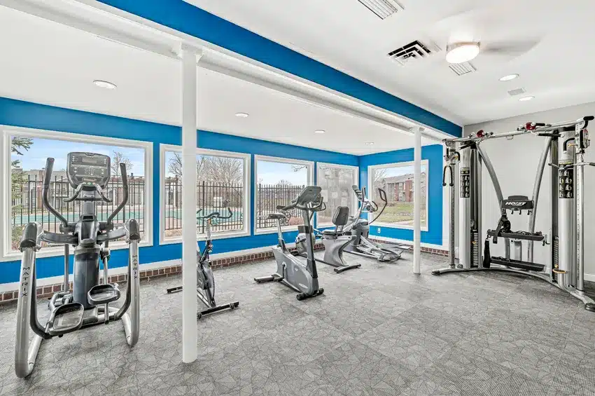 A gym room with cardio machines including an elliptical, stationary bikes, and a strength training machine, with large windows and blue accent walls.