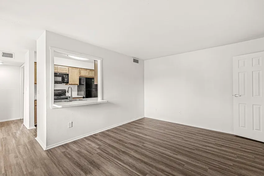 Unfurnished room with white walls, wood-style flooring, a closed white door, and a kitchen visible through a large interior window opening.
