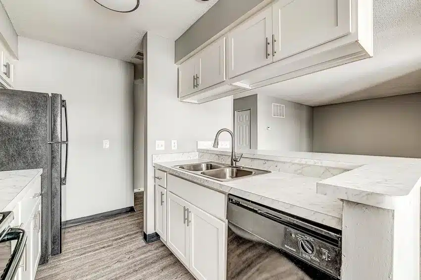 Modern kitchen with white cabinets, marble countertops, stainless steel sink, black dishwasher, and black appliances. Open layout with wood-style flooring and neutral-colored walls.