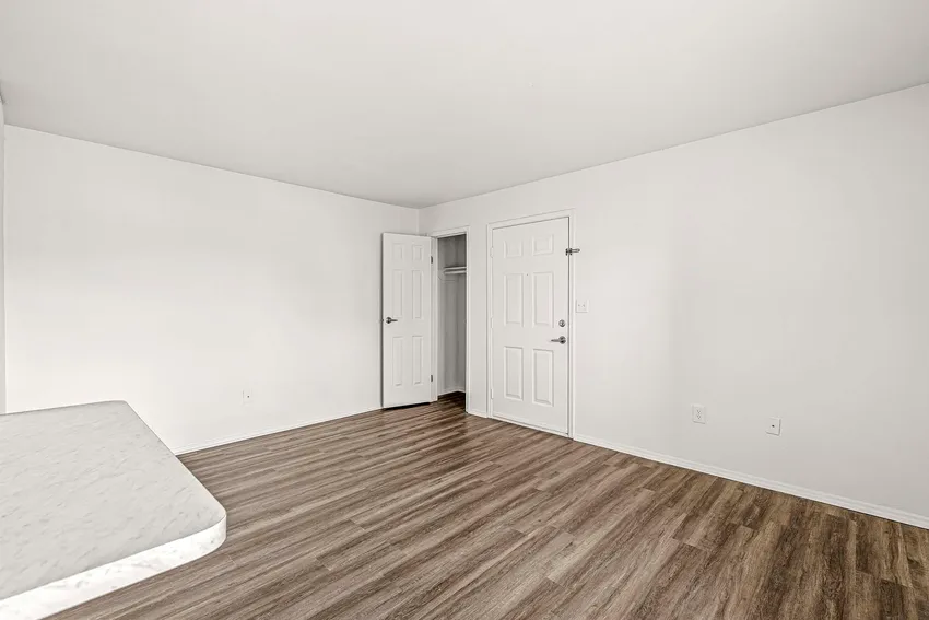 Empty room with white walls, wood-style flooring, a closet with an open door, and another closed door; part of a mattress is visible in the lower left corner.