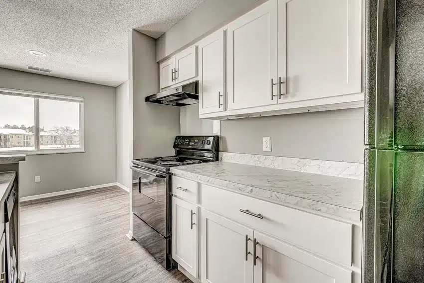 Modern kitchen with white cabinets, marble-style countertops, black stove, and a stainless steel refrigerator; window in the adjacent dining area lets in natural light.