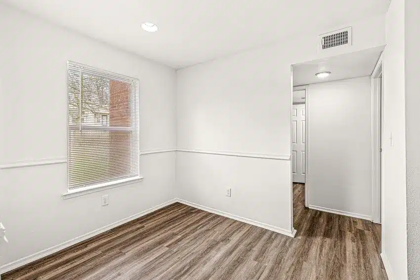 A small, empty room with white walls, a single window with blinds, wood-style flooring, and an open doorway leading to a hallway.