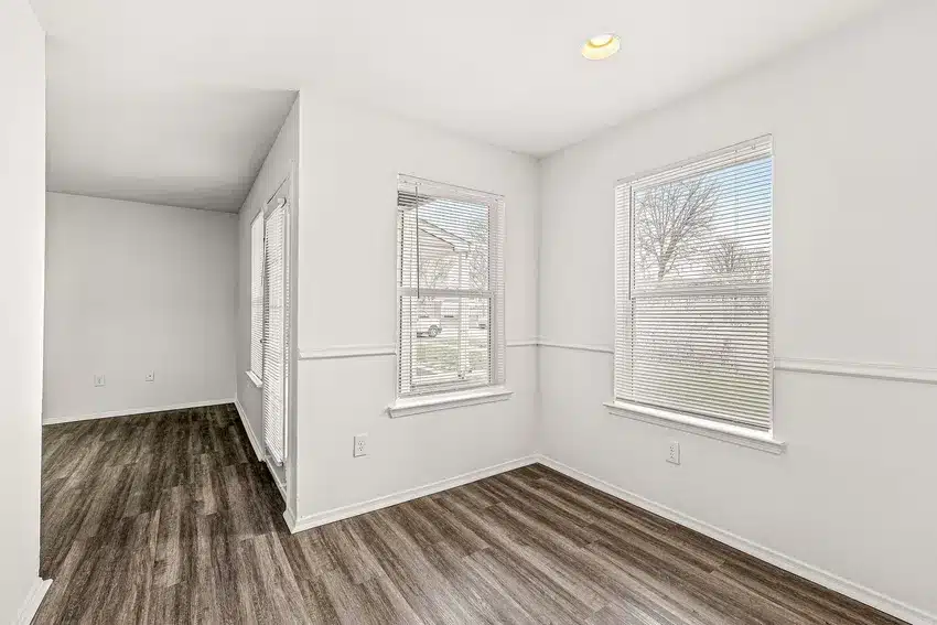 A bright, empty room with white walls, large windows with blinds, and dark wood flooring.