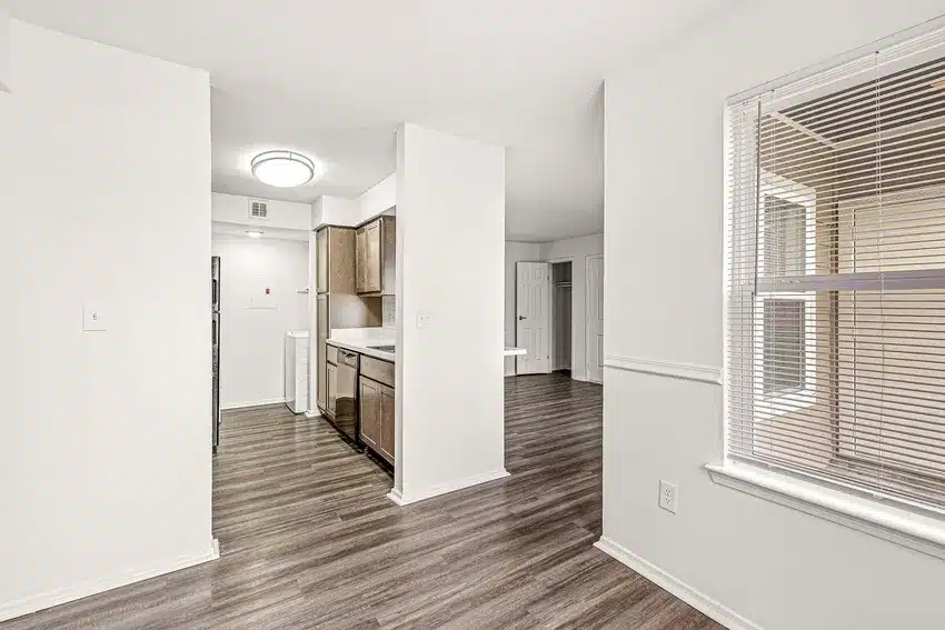 Modern apartment interior featuring a kitchen with wood cabinets, adjacent dining area, large window with blinds, and wood-look flooring throughout.