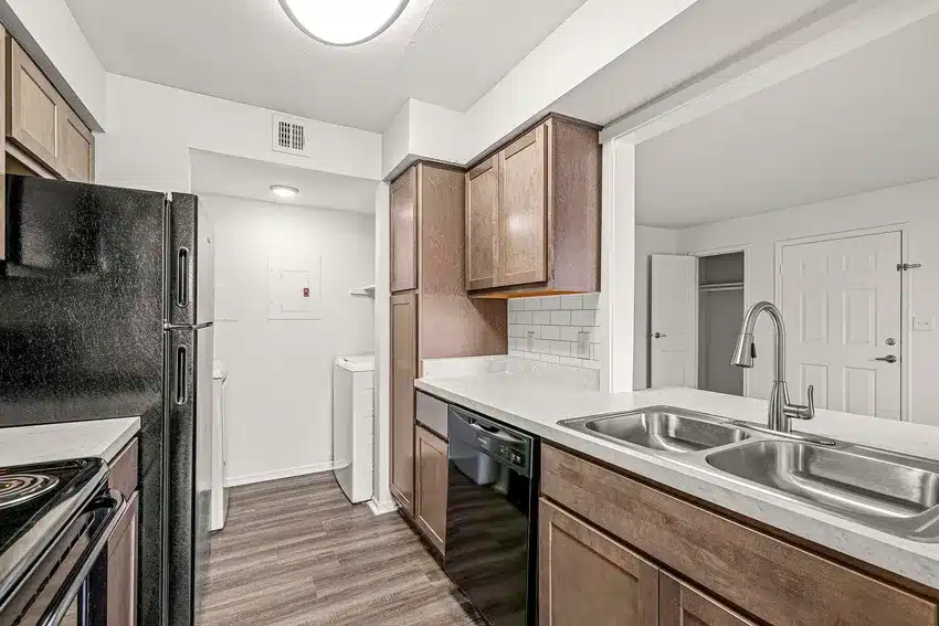Modern apartment kitchen with dark appliances, brown cabinets, white countertops, a double sink, and a view into a laundry area with a washer and dryer.