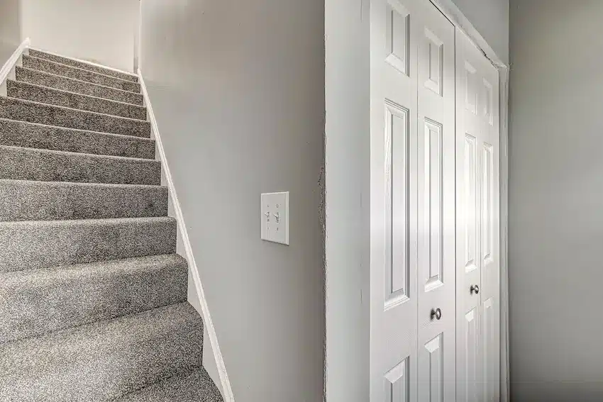Gray carpeted staircase next to a white closet with double doors in a hallway with light gray walls and a light switch on the wall.