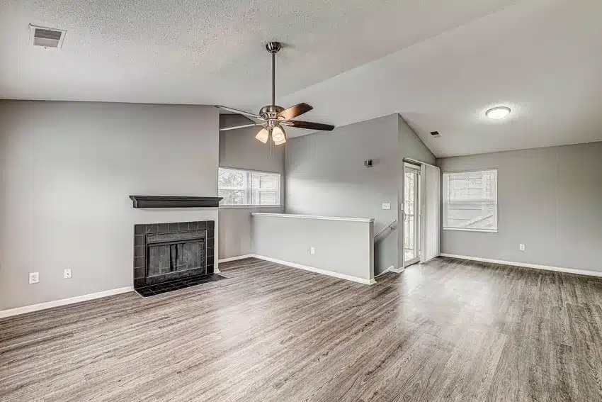 Empty living room with gray walls, wood-style flooring, a black fireplace, ceiling fan, and large window; adjacent to a dining area with a glass door and additional window.
