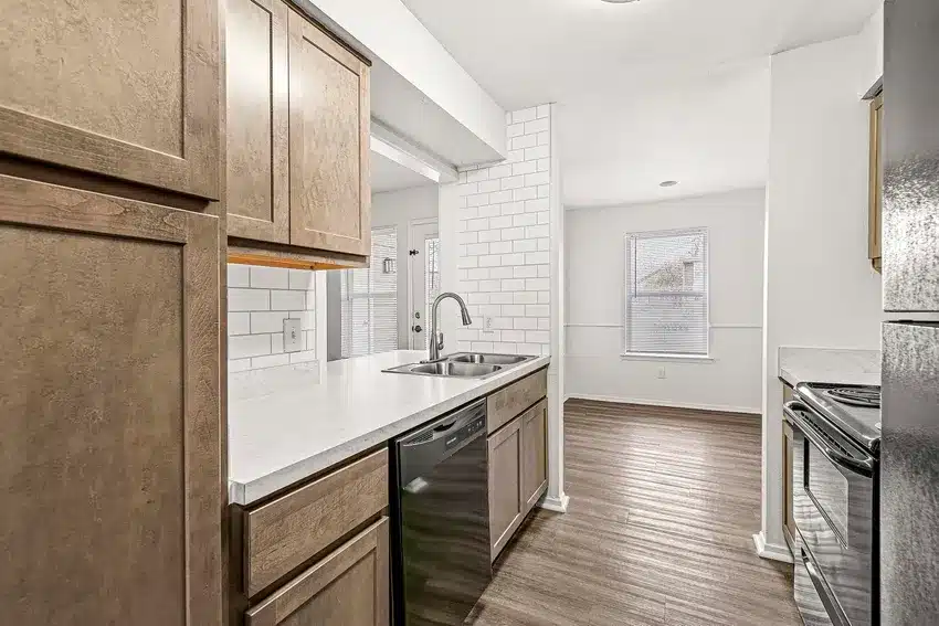 Modern kitchen with wood cabinets, white countertops, stainless steel sink, black appliances, and white subway tile backsplash, opening into a dining area with large windows and wood flooring.